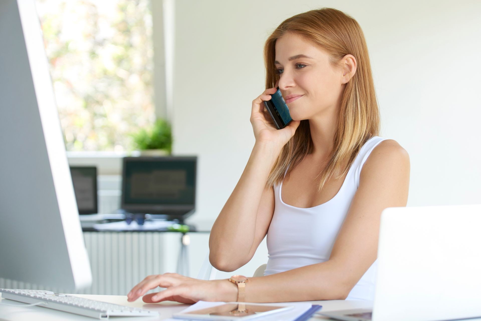 Beautiful young businesswoman talking with someone on her mobile phone while sitting in front of computer. 