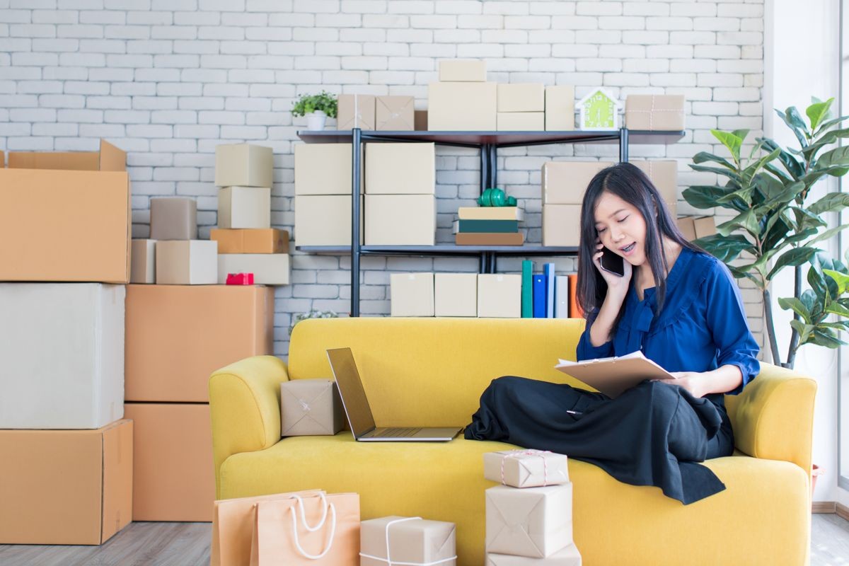 Young and beautiful Asian woman sitting among several boxes and calling on yellow sofa, working in the house office. Concept for home base business and startup ownership.